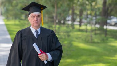 Diplomasını açık havada tutan mezuniyet elbisesi giymiş ciddi bir ihtiyar.