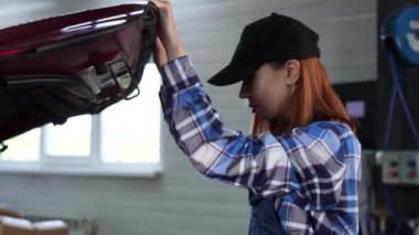 Kadın oto tamircisi araba servisinde arabanın kaputunun altına bakıyor.