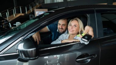 Kafkasyalı mutlu çift bir araba galerisinde arabada oturuyor. Bir kadın yeni bir arabanın anahtarlarını tutuyor.
