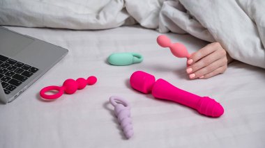 Yatakta yatarken dizüstü bilgisayarın yanında anal boncuk tutan bir kadın. Mastürbasyon için kişisel seks oyuncakları koleksiyonu
