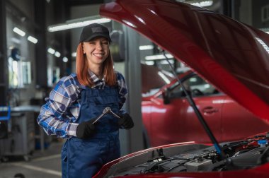 Kadın bir oto tamircisi arabanın açık kaputunda duruyor ve elinde bir tornavida tutuyor.