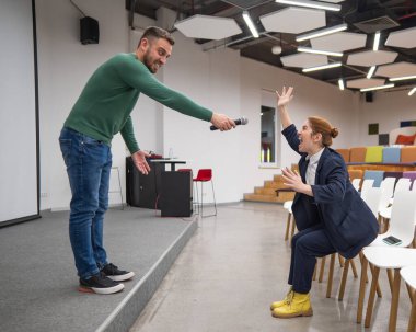 A red-haired Caucasian business woman sits in the front row in an empty conference room. Bearded man asks a question in the hall