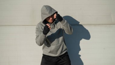 Kapüşonlu bir adam gri bir duvara karşı boks antrenmanı yapıyor.