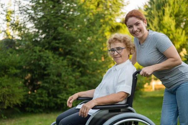 一位高加索妇女背着一位坐在轮椅上的老年母亲穿过公园 出去走走 — 图库照片