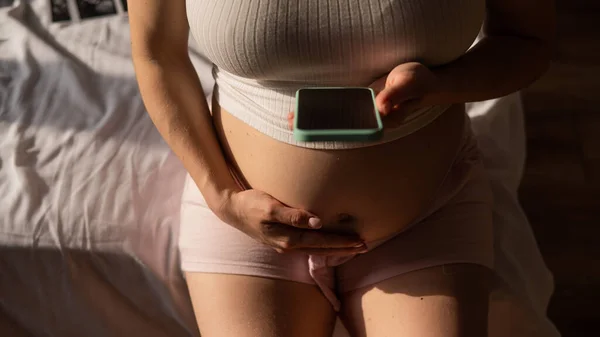 妊娠中の女性はベッドに座っている間スマートフォンを使用します 妊娠中の女性の腹のクローズアップ — ストック写真