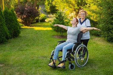 An elderly nurse walks with a middle-aged woman in a wheelchair through the park. The girl spread her arms outstretched like wings