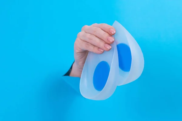 Woman holding orthopedic silicone heel pads. A womans hand sticks out of a hole in a cardboard blue background