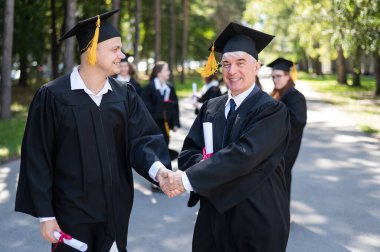 Bir grup mezun açık havada bornozla geziyor. Yaşlı bir adam ve genç bir adam diploma aldıkları için birbirlerini kutluyorlar.