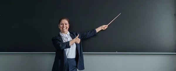 パンツ姿の赤髪の白人女性が親指を立てている 黒板にポインタで女性教師を笑顔 ワイドスクリーン — ストック写真
