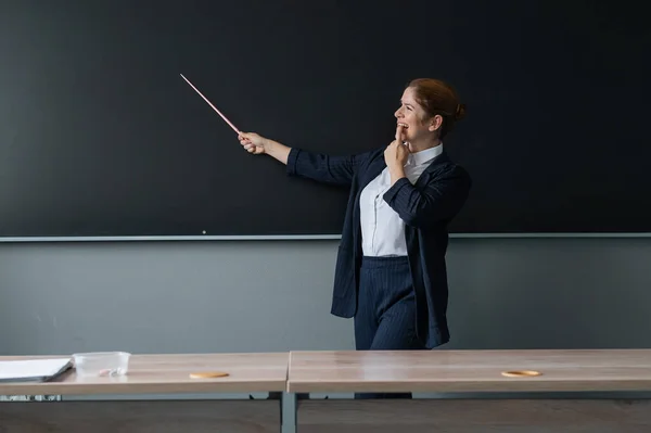Red-haired caucasian woman in a trouser suit. Thoughtful female teacher with a pointer at the blackboard