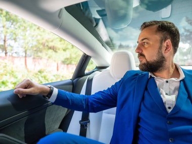 Mavi takım elbiseli beyaz bir adam arabanın arka koltuğunda otururken açık pencereden dışarı bakar. Business class yolcusu