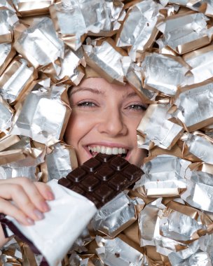 The face of a Caucasian woman surrounded by candy wrappers. The girl eats a bar of chocolate