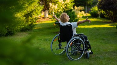 Rear view of an elderly woman spread her arms to the sides while sitting in a wheelchair on a walk outdoors