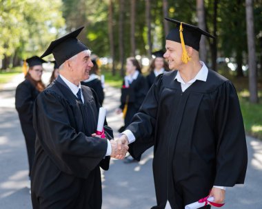 Bir grup mezun açık havada bornozla geziyor. Yaşlı bir adam ve genç bir adam diploma aldıkları için birbirlerini kutluyorlar.