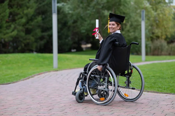 Stock image Happy caucasian woman in a wheelchair turns around and holds her diploma outdoors