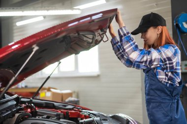 Kadın oto tamircisi araba servisinde bir arabanın kaputunu açıyor.