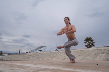 Beyaz bir adam, çıplak gövdesiyle deniz kıyısında wushu yapıyor.