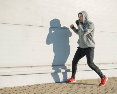 Kapüşonlu bir adam gri bir duvara karşı boks antrenmanı yapıyor.