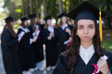 Mezuniyet elbisesi giymiş bir grup mutlu öğrenci dışarıda. Genç bir kızın elinde diploması var.