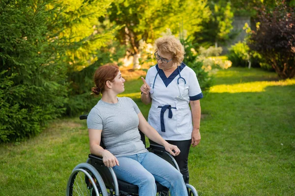 一位年长的女护士和一位坐在轮椅上的中年妇女在公园里散步 — 图库照片