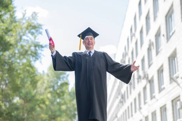 大学のローブを着た高齢者の白髪の男が腕を横に広げ 屋外で卒業証書を保持します — ストック写真