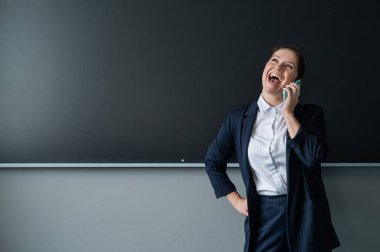 Beyaz kadın öğretmen, takım elbiseli. Karatahtanın önünde konuşurken telefonla konuşuyor.