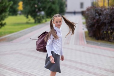 Üniformalı ve sırt çantalı mutlu bir beyaz kız okuldan sonra dışarı çıkıyor.