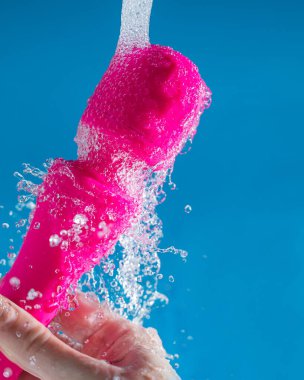 Bir kadın mavi arka planda akan suyun altında pembe bir vibratörü yıkıyor. Seks oyuncağı hijyen konsepti