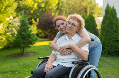 Beyaz bir kadın tekerlekli sandalyede oturan yaşlı bir anneye sarılıyor. Dışarı çık.