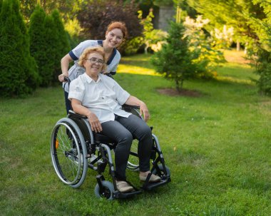 Beyaz kadın doktor yaşlı bir hastayla parkta tekerlekli sandalyede yürüyor. Hemşire dışarıda yürüyen yaşlı bir kadına eşlik ediyor.