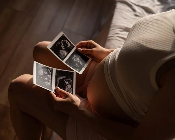 妊娠中の女性はベッドに座りながら超音波から写真を見ている 妊娠中の女性の腹のクローズアップ — ストック写真