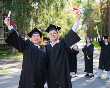 Bir grup mezun açık havada bornozla geziyor. Yaşlı bir adam ve genç bir adam diploma aldıkları için birbirlerini kutluyorlar.