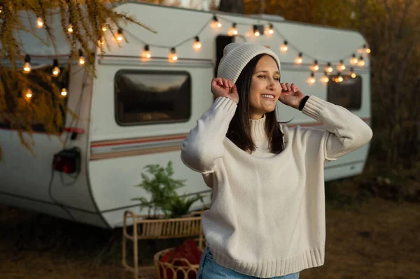 stock image A pensive Caucasian woman in a white knitted sweater and a hat is resting near a motorhome in a warm autumn. Travel by camper