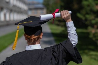 Mezuniyet elbisesi giymiş, elinde diplomasıyla üniversitenin yanında duran beyaz bir kadının arka görünüşü.