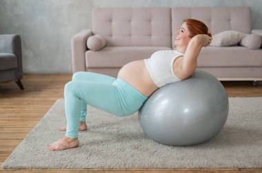 Hamile kızıl saçlı kadın evde fitball ile kol egzersizi yapıyor.