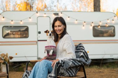 Beyaz kadın hasır bir sandalyede vagonun yanında oturuyor ve dışarıda Jack Russell Terrier 'ın köpeğine sarılıyor. Sonbaharda bir karavanla seyahat et.