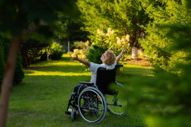 Rear view of an elderly woman spread her arms to the sides while sitting in a wheelchair on a walk outdoors