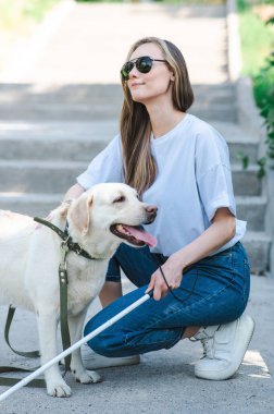 Kör genç sarışın parkta yürürken bir rehber köpeği okşuyor. Merdivenlerde değnekli bir kadın var.