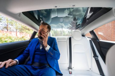 A caucasian man in a blue suit is talking on the phone in the back seat of a car. Business class passenger