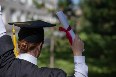 Mezuniyet elbisesi giymiş, elinde diplomasıyla üniversitenin yanında duran beyaz bir kadının arka görünüşü.