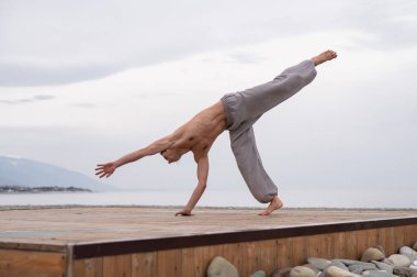 Beyaz bir adam, çıplak gövdesiyle deniz kıyısında wushu yapıyor.