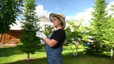Şapkalı ve tulumlu yaşlı bir kadının portresi bir bahçe aleti tutuyor. Video 360 derece