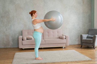 Hamile kızıl saçlı kadın evde fitball ile kol egzersizi yapıyor.