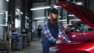 Otomobil tamircisi bir kadın, kollarını göğsüne bağlamış bir arabanın açık kaputunda duruyor.