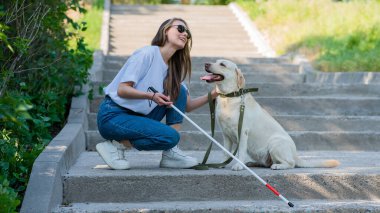 Kör genç sarışın parkta yürürken bir rehber köpeği okşuyor. Merdivenlerde değnekli bir kadın var.