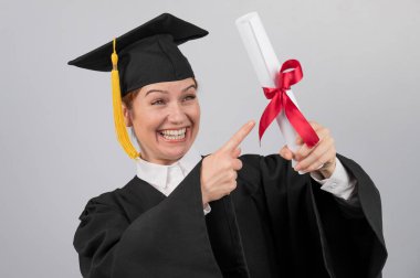 Mezuniyet elbisesi giymiş mutlu bir kadın diplomasını işaret ediyor.