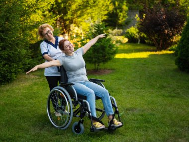 An elderly nurse walks with a middle-aged woman in a wheelchair through the park. The girl spread her arms outstretched like wings