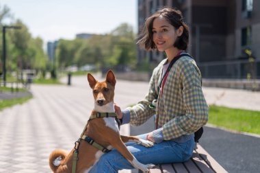 Genç ve güzel bir kadın açık havada bir köpekle bankta oturuyor. Havlamayan Afrika Basenji köpeği