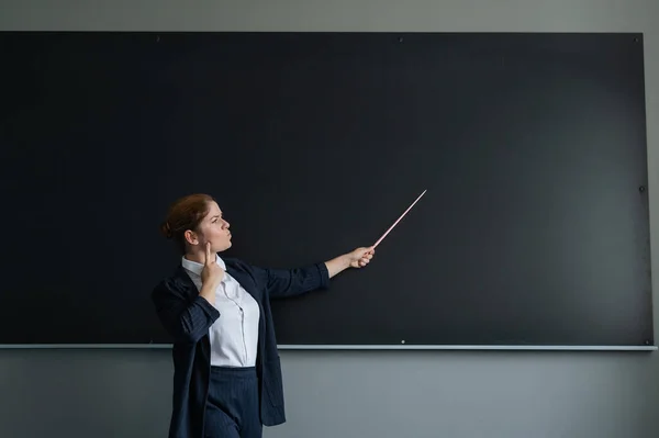ズボン姿の赤髪の白人女性 黒板にポインタを持つ思慮深い女性教師 — ストック写真