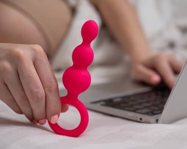 Yatakta yatarken dizüstü bilgisayarın yanında pembe anal boncuklar tutan kadın.